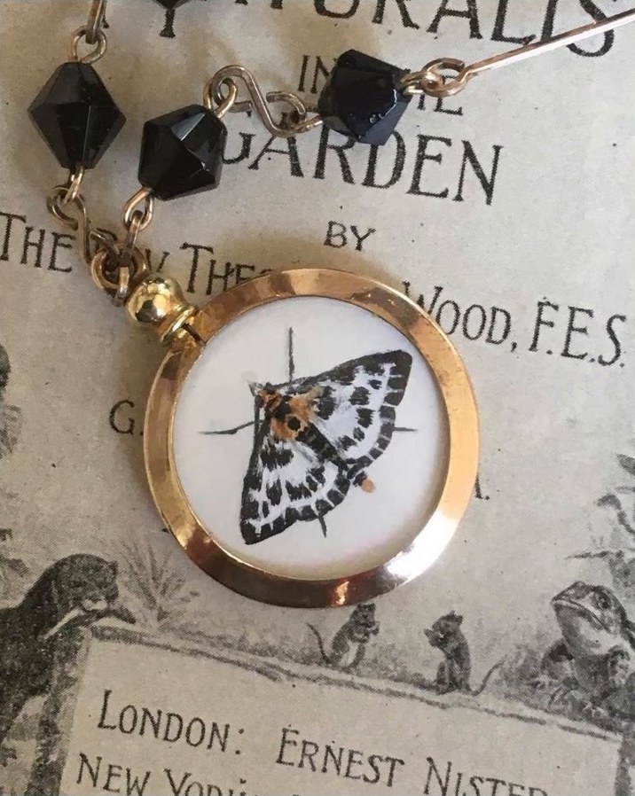 A commissioned painting of a black and white moth inside a gold locket on a jet bead chain, lying on an old natural history book